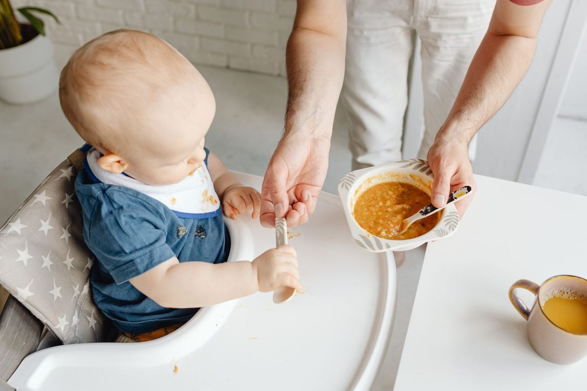 Baby som sitter støtt i barnestol og spiser puré med skje.